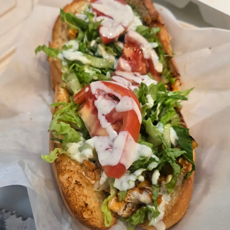 A close-up of a chicken sub sandwich with lettuce, tomatoes, and white sauce on a white paper.